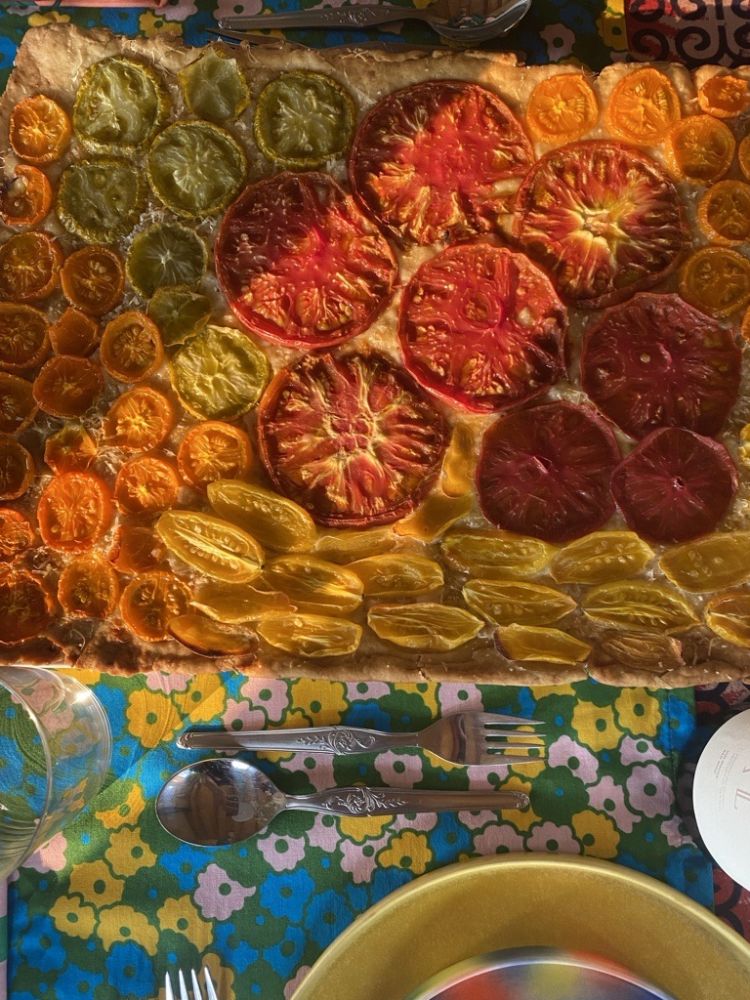 Tarte salée ou focaccia rectangulaire garnie de tranches de tomates multicolores (rouges, oranges, jaunes, vertes) déposée sur une nappe à motifs floraux colorés.