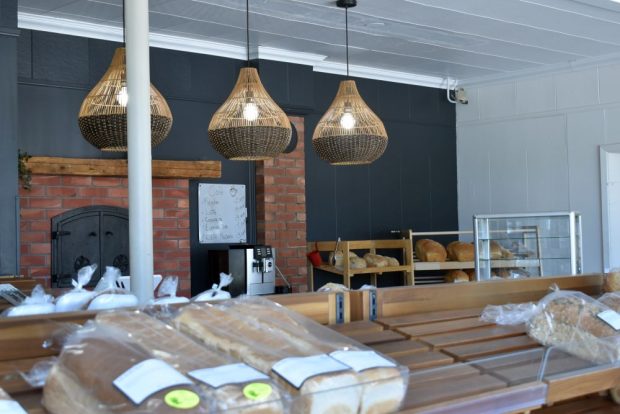 Photo couleur : Intérieur d'une boulangerie artisanale. Au premier plan, du pain emballé est exposé sur un présentoir en bois. À l'arrière-plan, un four en briques et des luminaires suspendus en osier contribuent à l'ambiance rustique et authentique du lieu.