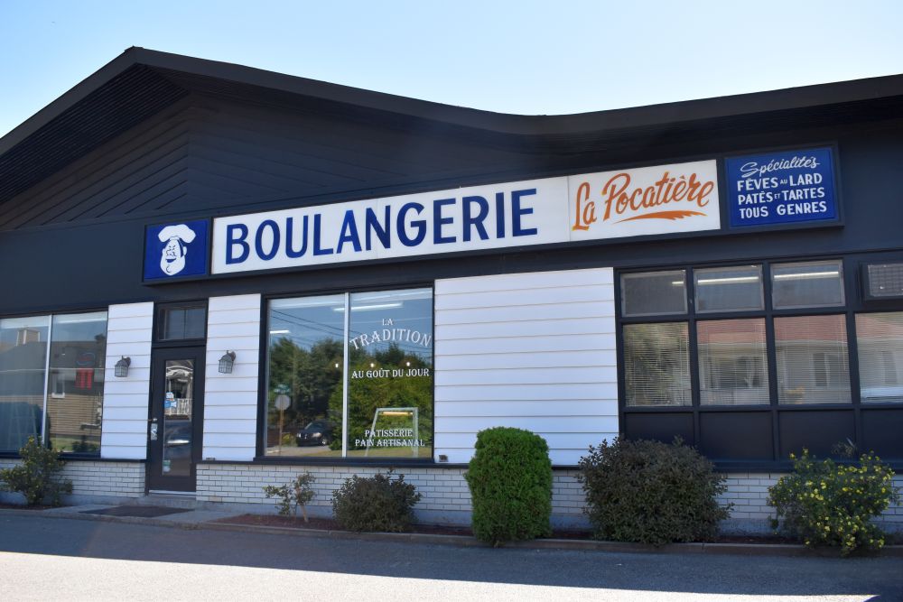 Photo couleur : Devanture de la "Boulangerie La Pocatière". L'enseigne claire, avec un logo de chef, indique les spécialités offertes comme les fèves au lard et les tartes. Le bâtiment sombre est agrémenté de parement blanc et de grandes fenêtres.