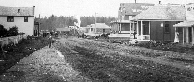 Photographie en noir et blanc d'une large route de terre bordée, de part et d'autre, de bâtiments. On voit, à droite, deux personnes, chacune debout à l'extérieur de l'entrée d’un bâtiment, et à l'arrière, sur la route, un groupe de personnes non loin d'une vieille automobile.