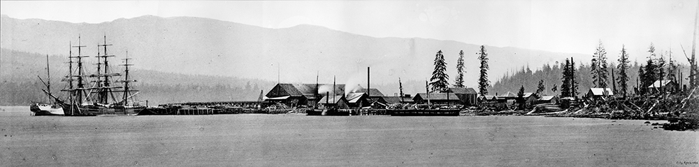 Une photographie en noir et blanc de la scierie Hastings en 1872. La scierie est située le long du rivage et est entourée de plusieurs petits bâtiments. Un navire européen est amarré au port, du côté gauche de l’image. 