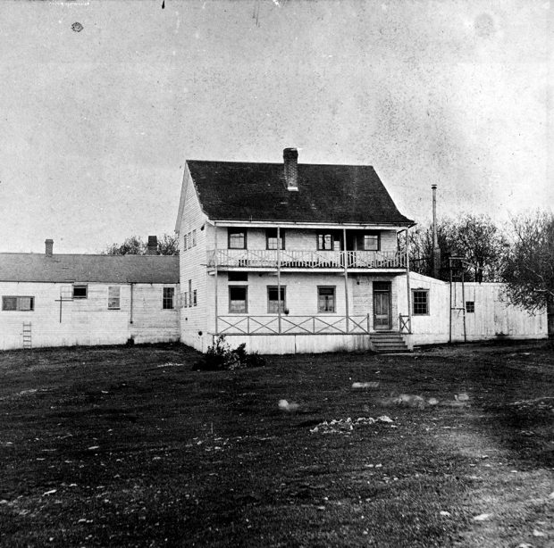 Une photo en noir et blanc de l’asile d’aliénés de Victoria. Le bâtiment principal, qui a deux étages, a un perron au rez-de-chaussée et un balcon au deuxième étage. Il y a un long bâtiment d’un étage situé derrière l’édifice principal.