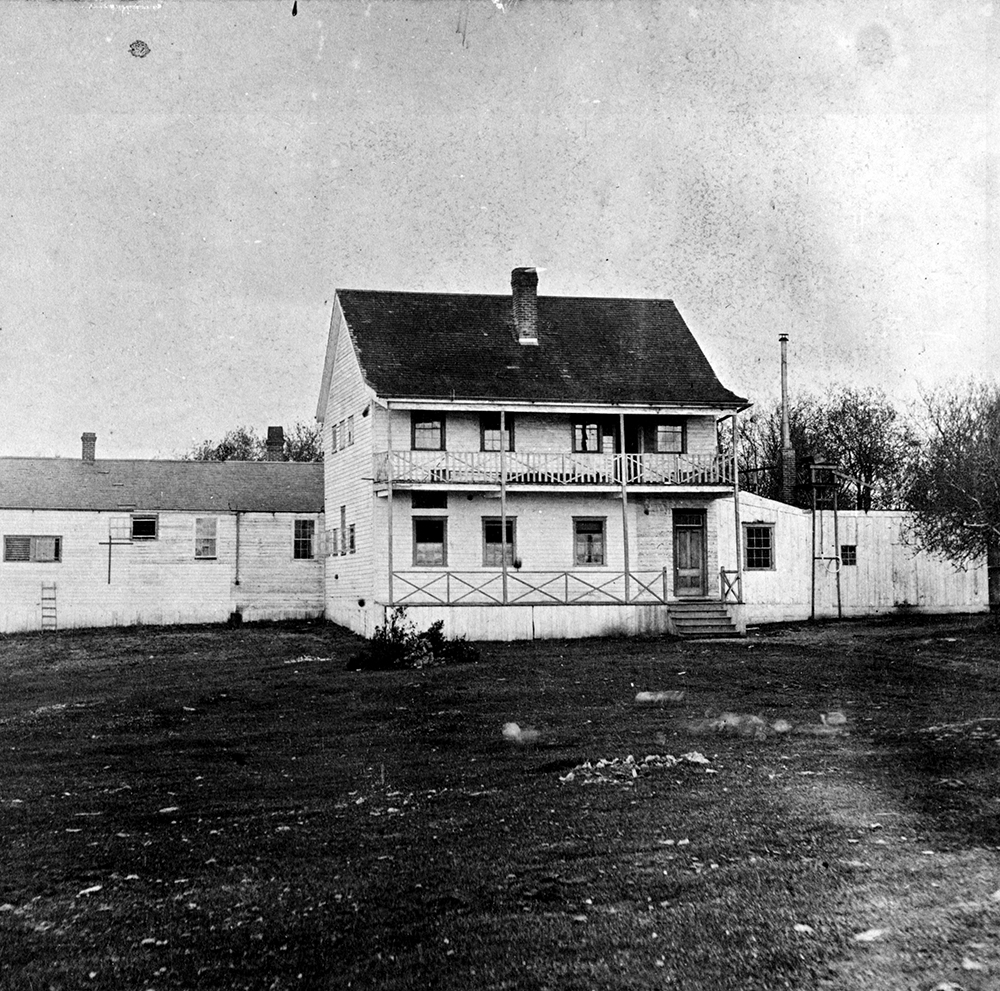 Une photo en noir et blanc de l’asile d’aliénés de Victoria. Le bâtiment principal, qui a deux étages, a un perron au rez-de-chaussée et un balcon au deuxième étage. Il y a un long bâtiment d’un étage situé derrière l’édifice principal.