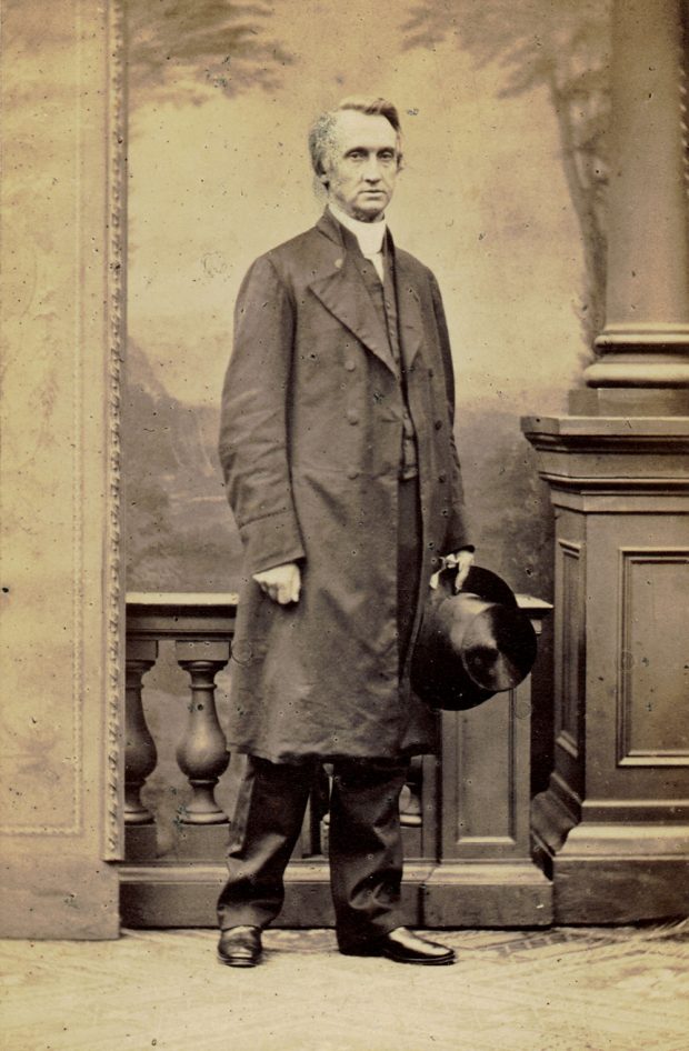 Une photographie en noir et blanc (réalisé en studio) de l’évêque George Hills vêtu d’un long manteau. Il tient un chapeau haut de forme à la main gauche et a le bras droit le long du corps, poing fermé.