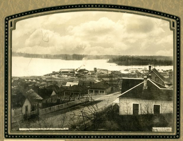 Une photographie monochrome aux tons sépia, prise en plongée, de New Westminster; on y voit l’usine de rabotage appelée Royal City Planing Mills sur la rive du fleuve Fraser. 