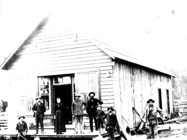Photographie en noir et blanc de six hommes, une femme et un garçon, debout devant le magasin de Shortreed. Le magasin est un grand bâtiment de bois avec une terrasse menant à la porte de la façade. Il y a une fenêtre de chaque côté de cette porte, et deux autres portes visibles à l’arrière-plan à droite, sur le côté du bâtiment.