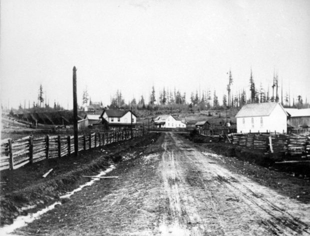 Photographie en noir et blanc de Murray’s Corners à partir de la route Old Yale, à Langley. La route monte une colline et est bordée, des deux côtés, de clôtures de bois. Il y a quatre grands bâtiments en haut de la colline.