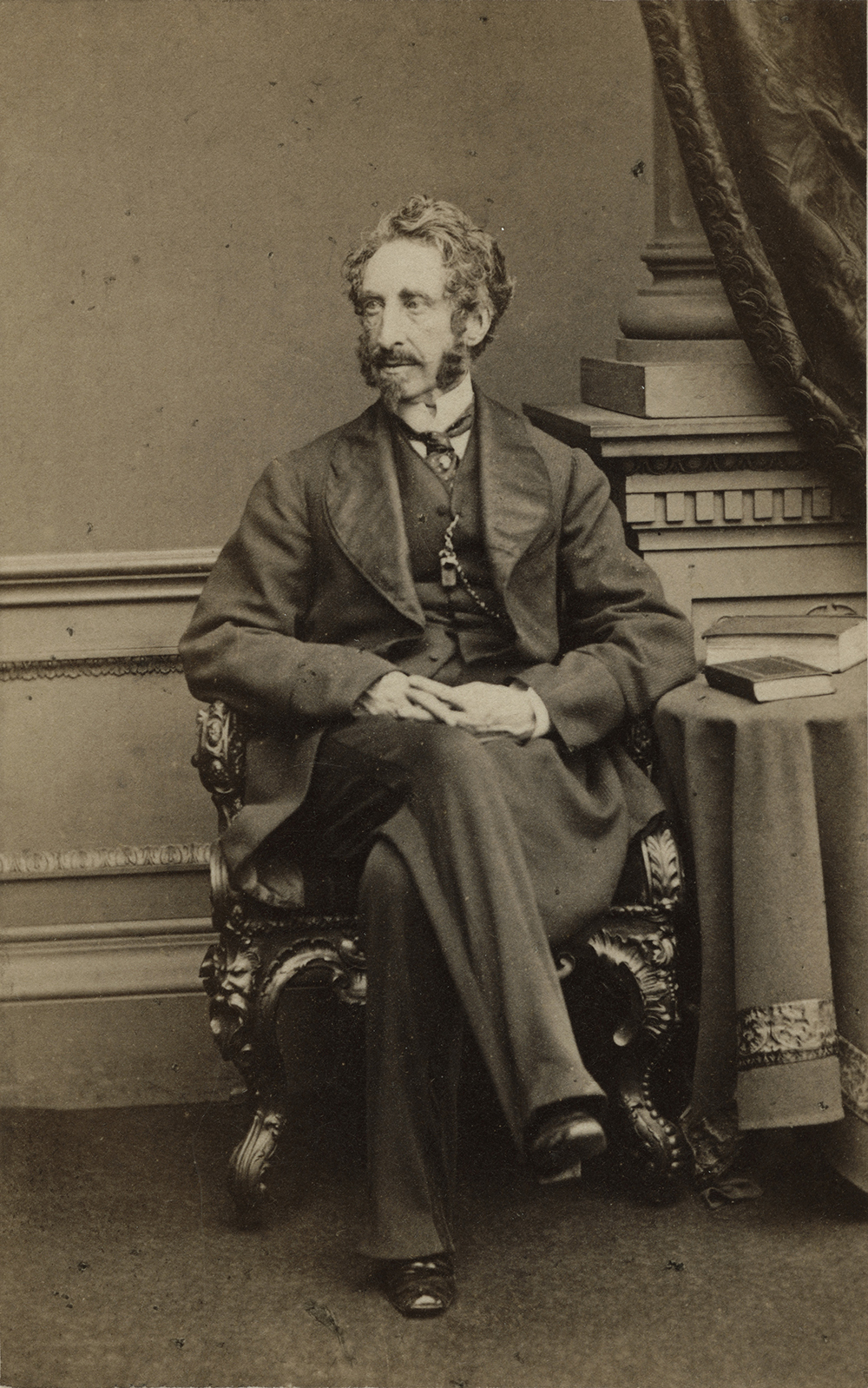 Portrait photo en noir et blanc (réalisé en studio) de Sir Edward Bulwer-Lytton, assis sur une chaise très ornée près d’une petite table où se trouvent deux livres. 