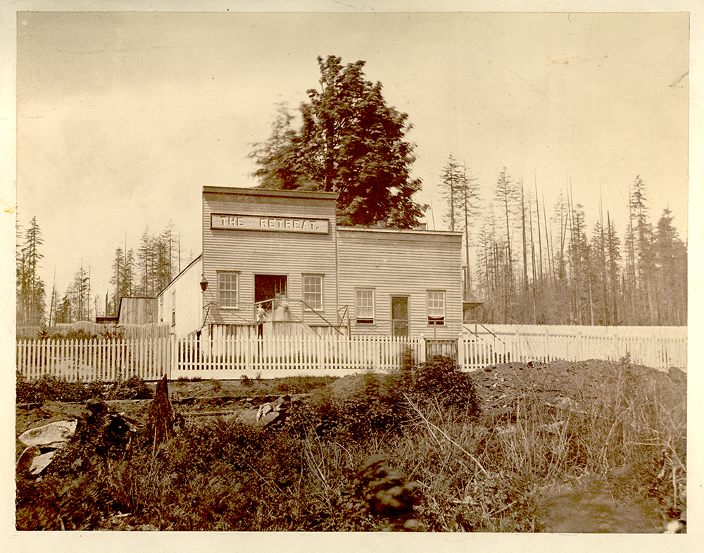 Une photographie monochrome aux tons sépia, prise autour de 1865, d’un bâtiment à deux devantures. À gauche, on voit un saloon qui a une fausse façade plus haute portant une enseigne où l’on peut lire « The Retreat ». Chaque devanture a sa propre porte et une fenêtre de part et d’autre de la porte. Il y a une clôture en lattis devant le bâtiment et une forêt à l’arrière-plan.  