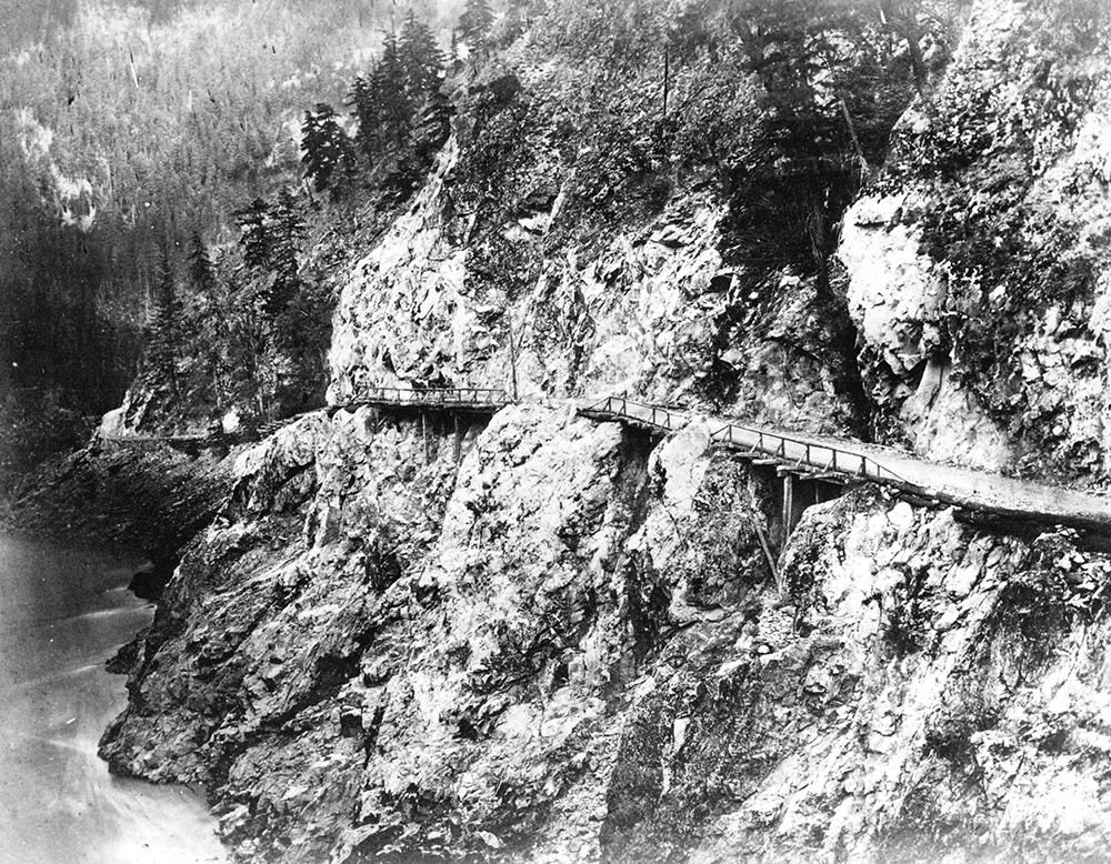 Une photographie en noir et blanc d’une étroite route accessible aux charrettes construite le long d’une falaise rocheuse surplombant une rivière. Certaines sections de la route sont des ponts, bordés du côté extérieur d’un garde-fou en bois.