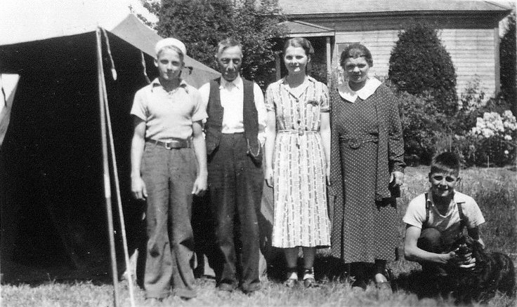 Photographie en noir et blanc d'un groupe composé de deux jeunes filles, deux jeunes garçons et un homme âgé près d'une tente, devant une maison. Tous sont debout, excepté l'un des garçons, à droite, qui est accroupi et caresse un chien.