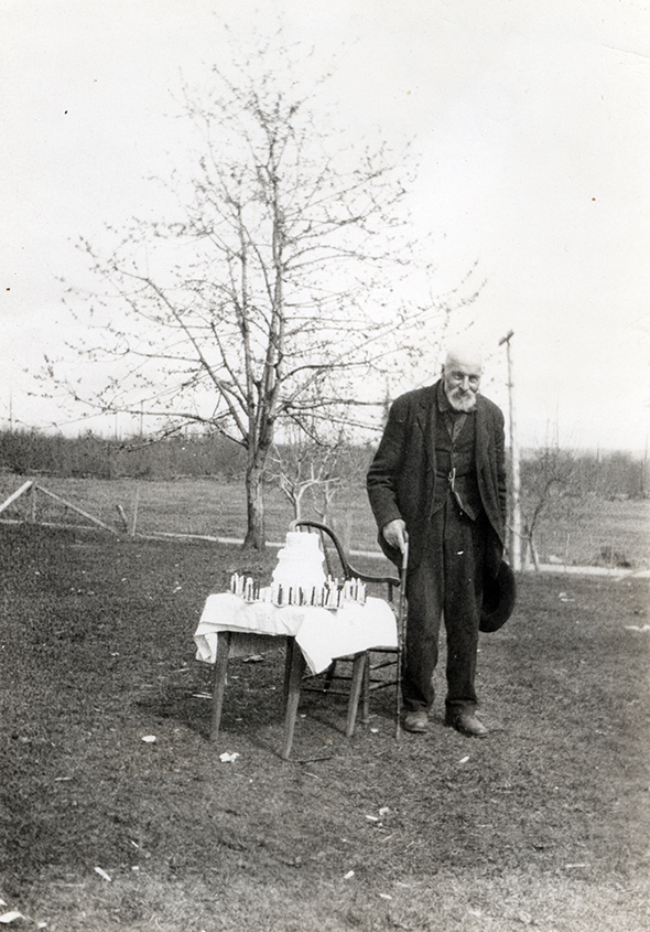 Photographie monochrome aux tons sépia d'un homme âgé debout près d'une table de bois sur laquelle est posé un gâteau à trois étages.
