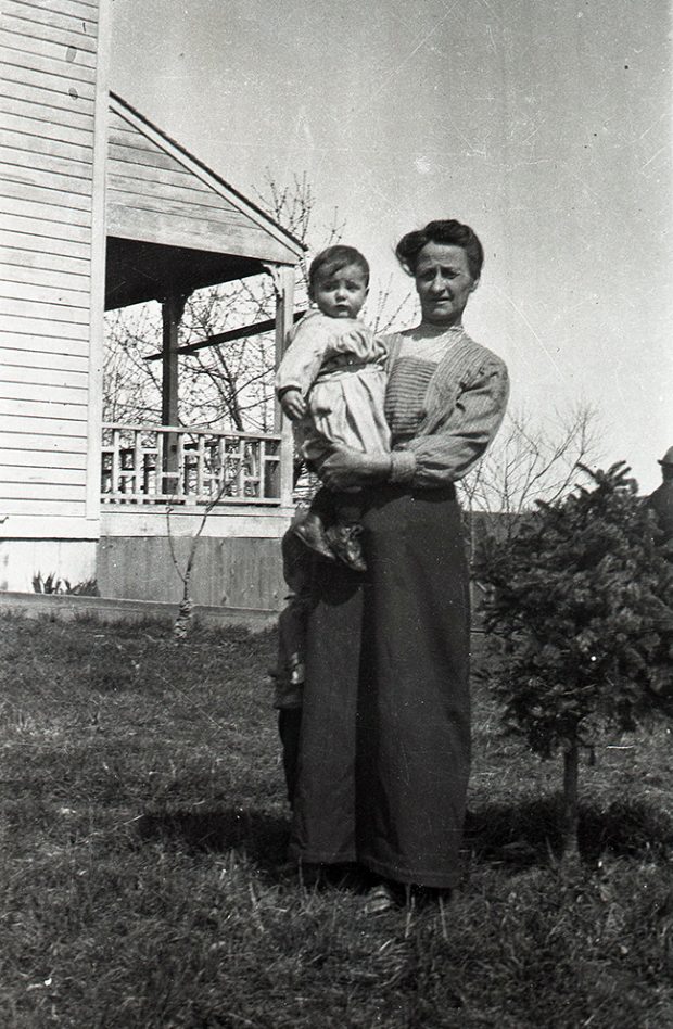 Photographie en noir et blanc d'une femme tenant un petit garçon dans ses bras, debout sur la pelouse près d’une maison.