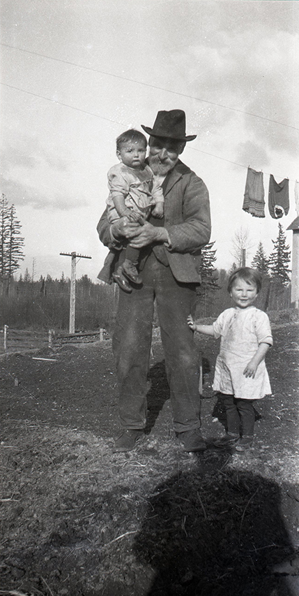 Photographie en noir et blanc d'un homme tenant un petit garçon dans ses bras en compagnie d’une petite fille, debout à côté de lui. La petite fille a une main appuyée sur la jambe de l'homme. À l'arrière-plan, du côté droit de l'image, une corde à linge d'où pendent quelques vêtements.