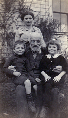 Photographie monochrome aux tons sépia d'un homme assis sur une chaise, avec un petit garçon et une petite fille sur ses genoux. Une femme est debout derrière eux, et on voit un bâtiment à l'arrière-plan.