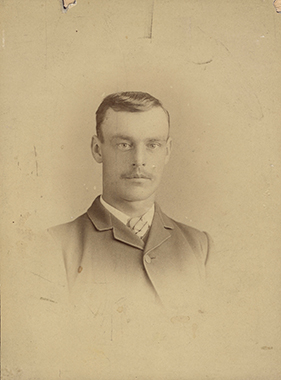 Photographie monochrome aux tons sépia (portrait réalisé en studio) d'un jeune homme moustachu.