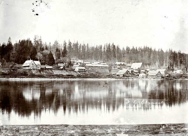 Photographie en noir et blanc d'une petite colonie établie près d'un fleuve. La colonie est constituée d'un groupe de petits bâtiments entourée, à l'arrière, d'une dense forêt.