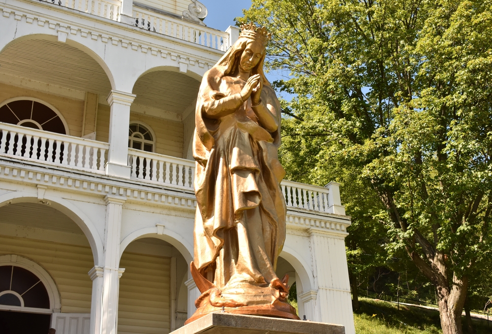 Photographie en couleurs d’une statue dorée de grand format représentant la Vierge Marie. La Vierge porte une couronne et se tient debout sur un dragon, les mains jointes devant elle. La statue est posée sur un socle et, à l’arrière-plan, on aperçoit un bâtiment à plusieurs étages.