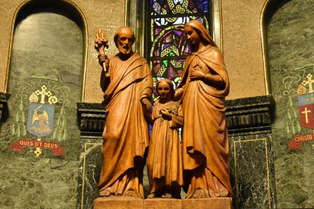 Photographie en couleurs d’une grande sculpture de bois représentant, de gauche à droite, Joseph, Jésus et Marie. La sculpture se trouve sur un socle dans une chapelle sombre et richement décorée.
