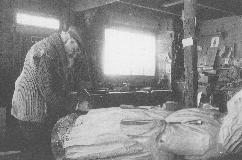 Photographie d’archives en noir et blanc. Un homme sculpte le pied d’une grande statue en bois à l’intérieur d’un atelier où sont éparpillés croquis et outils servant à travailler le bois.