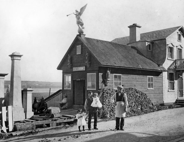 Photographie d’archives en noir et blanc. Deux hommes posent debout devant un édifice composé de deux sections. La partie de gauche, à un étage, porte l’inscription « L. JOBIN » au-dessus de la porte. La partie de droite, à deux étages, est une maison. Un des hommes est accompagné de deux jeunes enfants.