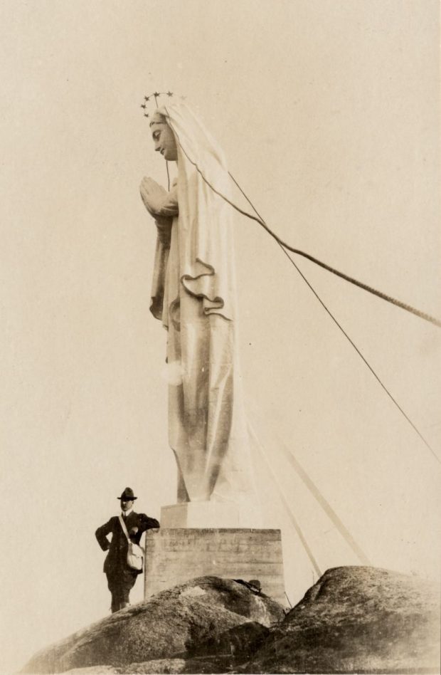 Photographie d’archives en noir et blanc. Un homme est accoudé au pied d’une statue faisant plusieurs fois sa hauteur. La sculpture blanche représente la Vierge Marie couronnée d’étoiles, les mains jointes devant elle. La statue est posée sur un socle qui est lui-même placé sur le roc.
