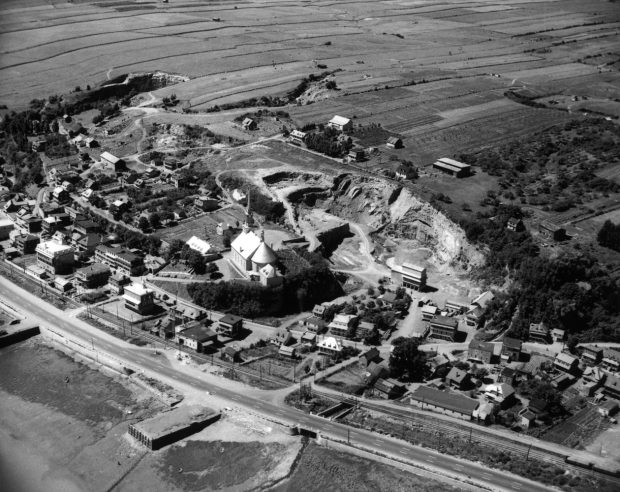 Photographie d’archives en noir et blanc. La municipalité de Château-Richer se déploie autour de l’église, qui se trouve au centre de l’image. Outre des maisons, on aperçoit une carrière de pierre, des champs cultivés, un chemin de fer et le boulevard Sainte-Anne.