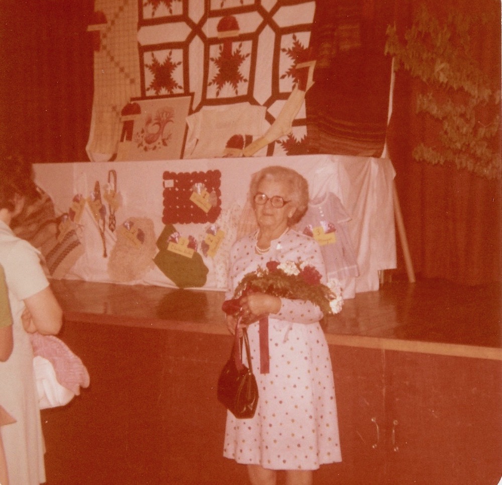 Photographie couleur d’Amanda Gagnon Jalbert tenant un bouquet de fleurs. Elle porte une robe blanche à motif imprimé et tient un sac à main. Derrière elle, sur une scène, se trouve une table où sont exposées des pièces d’artisanat textile.