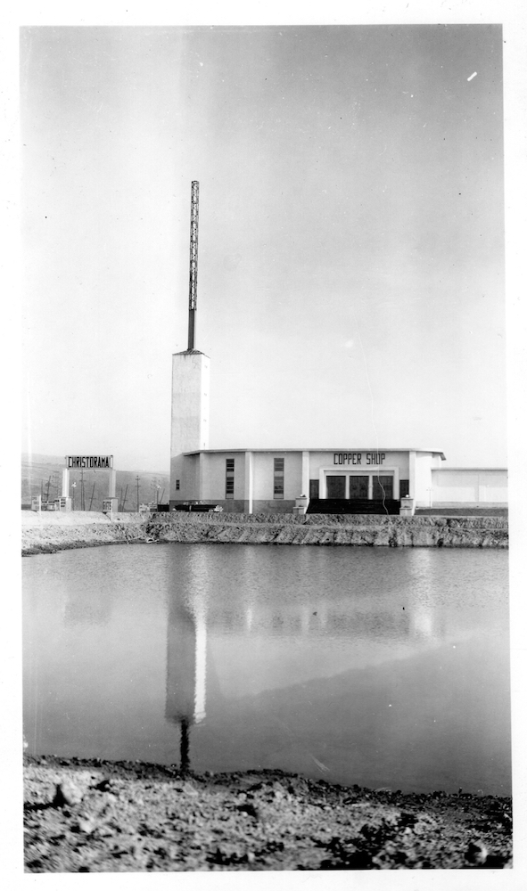 Photographie d’archives en noir et blanc montrant un bâtiment moderne blanc de deux étages sur le bord d’une route, avec une cheminée à l’extrémité gauche. Devant, on voit la réflexion du bâtiment dans un plan d’eau. À gauche, sur une grande enseigne, on peut lire « Christorama ». Il est aussi écrit « Copper Shop » ou « atelier de cuivre » sur le bâtiment.