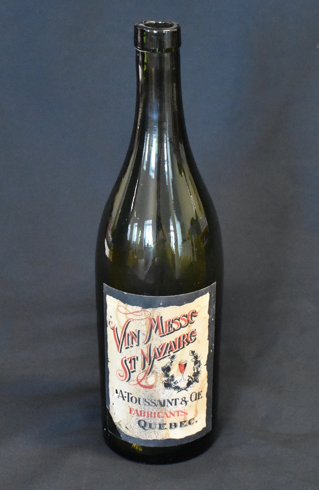 Photographie couleur sur fond noir d’une bouteille de vin à goulot allongé. La bouteille porte une étiquette sur laquelle on peut lire « Vin de messe St Nazaire A. Toussaint & Cie. Fabricants, Québec ».