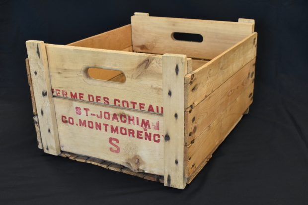 Photographie couleur d’une caisse faite de lattes de bois portant, écrite en rouge, l’inscription « Ferme des coteaux St-Joachim Co. Montmorency S » sur le côté court faisant face à la caméra.