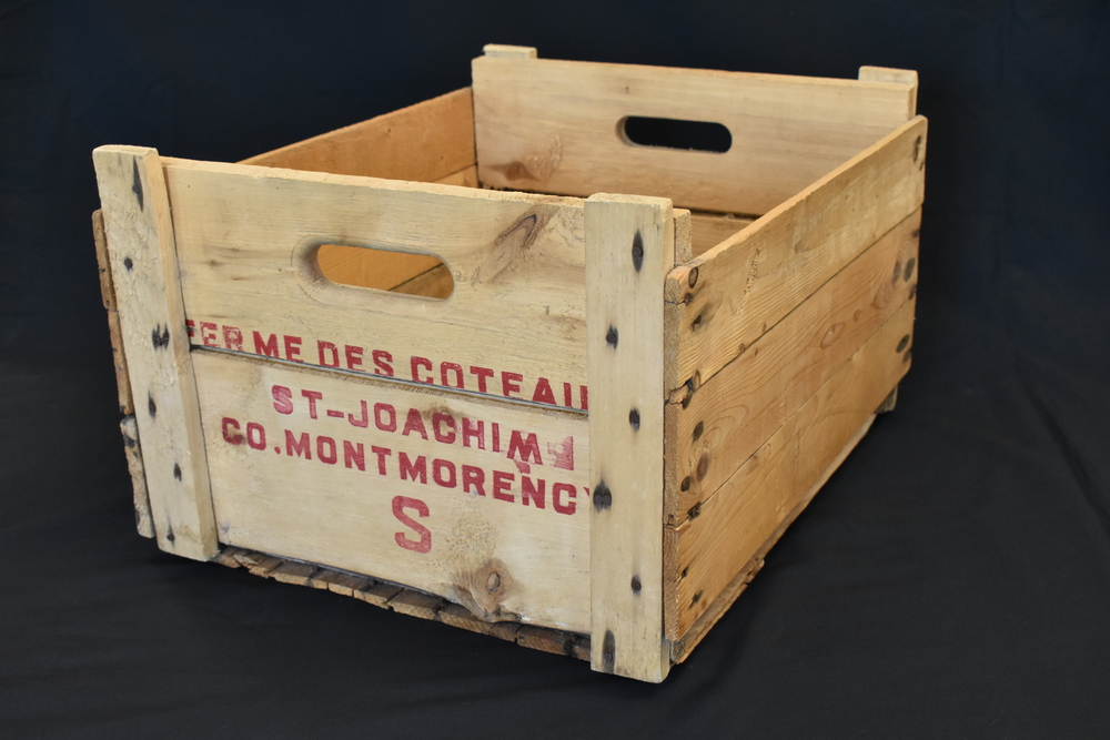Photographie couleur d’une caisse faite de lattes de bois portant, écrite en rouge, l’inscription « Ferme des coteaux St-Joachim Co. Montmorency S » sur le côté court faisant face à la caméra.