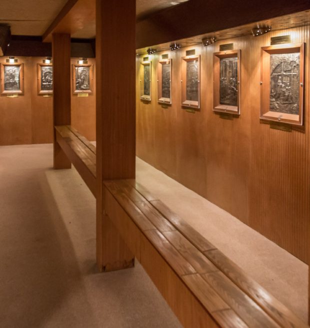 Photographie couleur montrant une salle aux murs de bois où est exposée une série de tableaux en argent repoussé. Chaque tableau est surmonté d’un petit écriteau descriptif. Un long banc traversé d’une colonne se trouve au centre de la pièce.