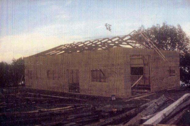 Photographie d’archives en couleurs d’un bâtiment en construction. On voit les murs en bois ainsi que la charpente du toit, également en bois. Sur le sol, sur l’avant et sur le côté du bâtiment, se trouvent des planches de bois et des troncs d’arbres.