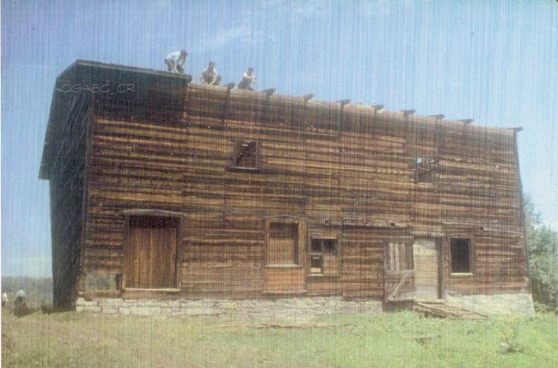 Photographie d’archives en couleurs d’une grange en bois avec des fondations en pierre. On y voit deux fenêtres à l’étage supérieur ainsi que deux portes et trois fenêtres à l’étage inférieur. Trois hommes travaillent à défaire la toiture.