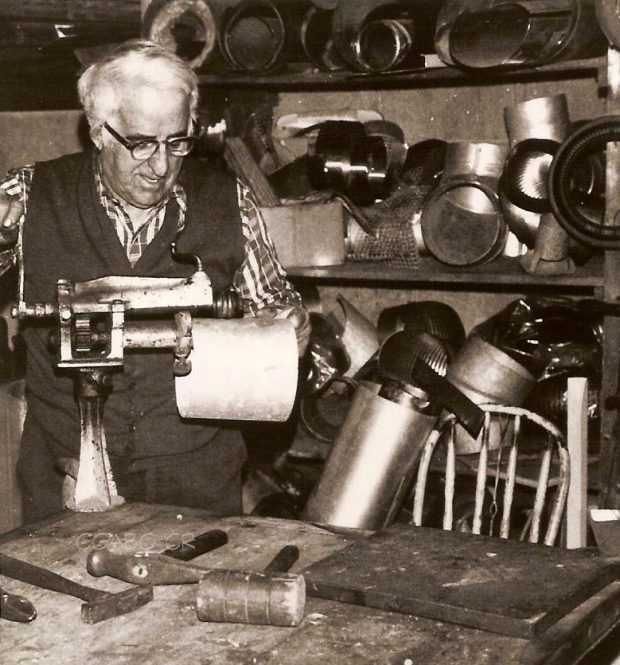 Photographie d’archives en noir et blanc montrant Joseph Drouin au travail dans son atelier de ferblanterie. Il actionne une machine de la main droite, tandis qu’il tient de la main gauche l’extrémité de l’objet en cours de fabrication. Des outils tels des marteaux et un maillet se trouvent sur une table devant lui. Les étagères à l’arrière sont jonchées de divers objets en métal.