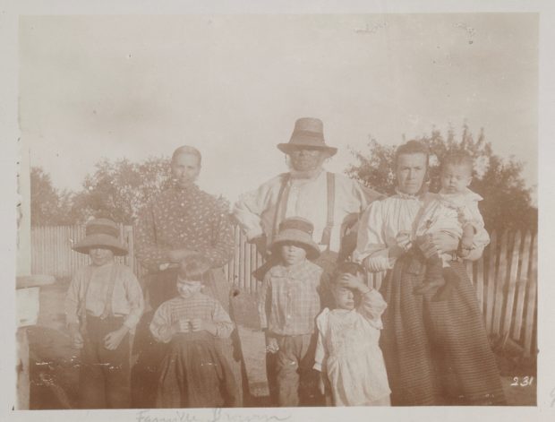 Photographie d’archives en noir et blanc d’une famille se tenant debout à l’extérieur, devant une clôture et des arbres. Coiffé d’un chapeau, Hugh Brown se tient au centre; de part et d’autre, on aperçoit deux femmes. La femme de droite tient un bambin dans ses bras. Quatre jeunes enfants se trouvent au premier plan, devant les adultes.