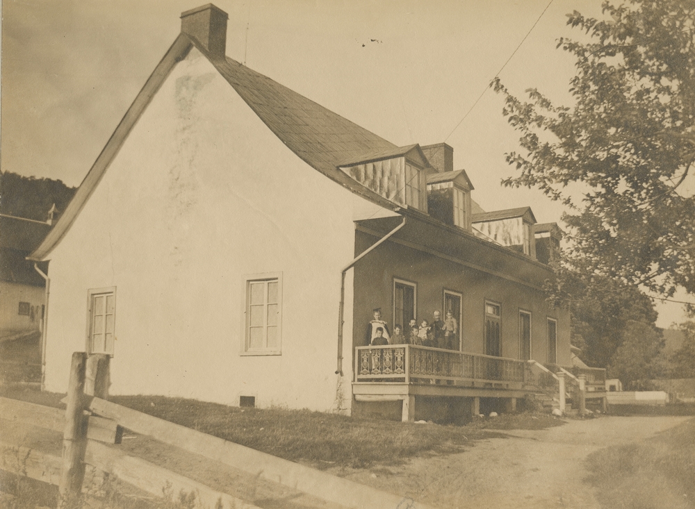 Photographie d’archives en noir et blanc du côté d’une maison blanche au toit à deux versants avec une galerie à l’avant. La maison comporte deux cheminées et quatre pignons. Sur la façade, quatre fenêtres, deux de chaque côté, entourent la porte. Sur la galerie, on voit une famille comptant sept enfants qui pose pour la photographie. On entrevoit un arbre et une allée devant la maison.