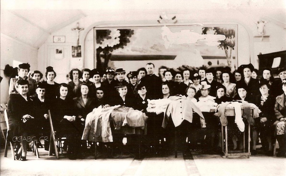 Photographie d’archives en noir et blanc où un groupe de femmes alignées sur deux rangs posent pour la caméra dans une grande salle. Le premier rang est composé de femmes assises, tandis que celles du deuxième rang sont debout. Un seul homme, un abbé, se trouve au centre. Des tables placées à l’avant du groupe montrent des pièces d’artisanat textile.