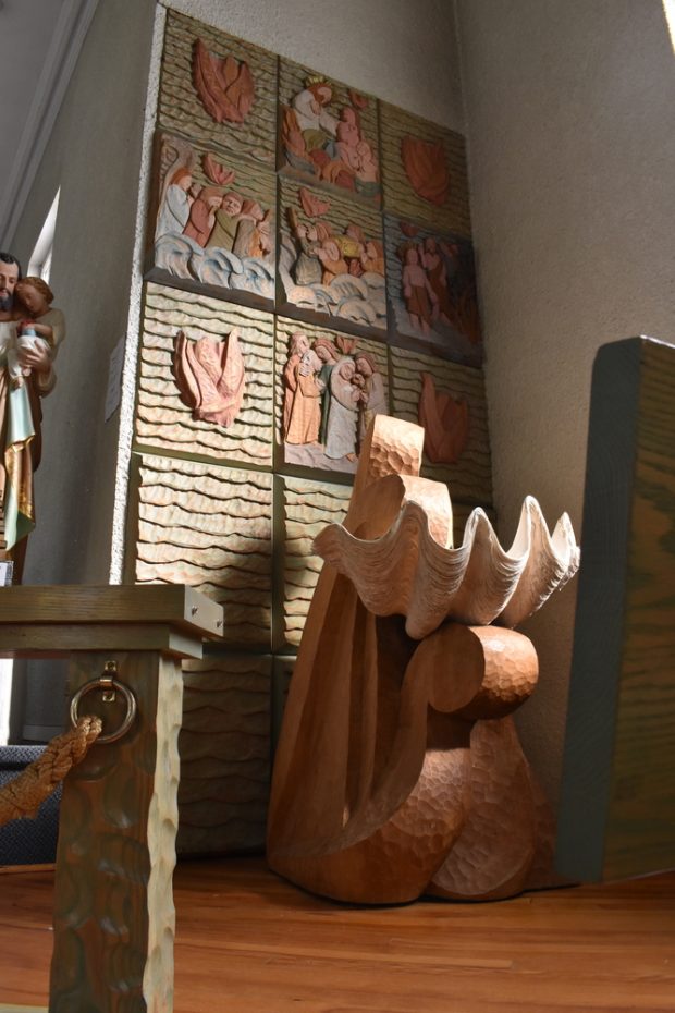 Photographie couleur de l’intérieur d’une église. Les fonts baptismaux, au centre, sont composés d’un immense coquillage et leur base présente des motifs de vagues. Sur le mur à l’arrière, les tableaux sculptés dans le bois et peints illustrent des scènes bibliques où l’eau et les vagues constituent un motif central.