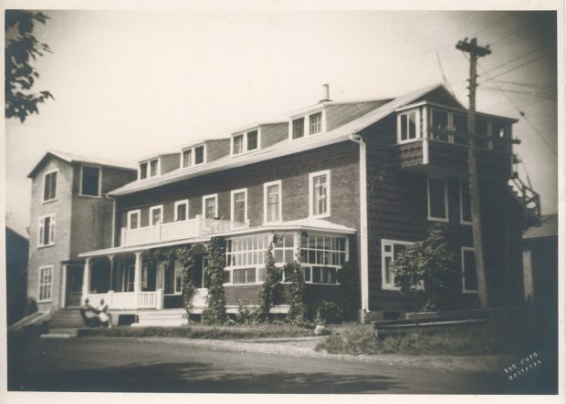 Photographie d’archives en noir et blanc montrant un hôtel à trois étages. Le toit compte deux versants. La façade principale comprend un balcon et une véranda. Le premier étage inclut un solarium.