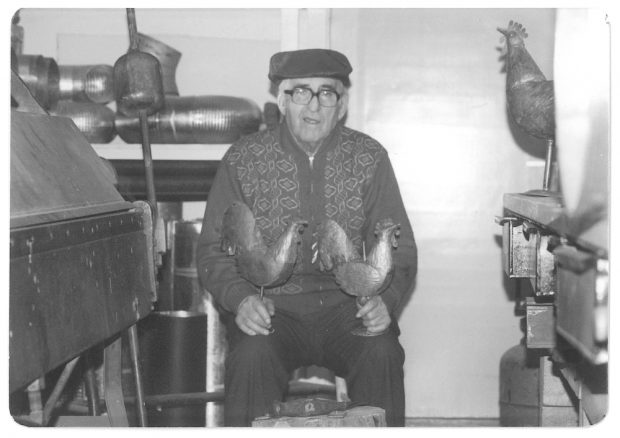 Photographie d’archives en noir et blanc montrant Joseph Drouin, âgé et assis dans son atelier, tenant un coq en cuivre sur chacun de ses genoux. Des morceaux de tuyaux en métal sont visibles sur une étagère derrière son épaule, à gauche, et on aperçoit un coq en cuivre plus grand à sa droite, sur un établi.