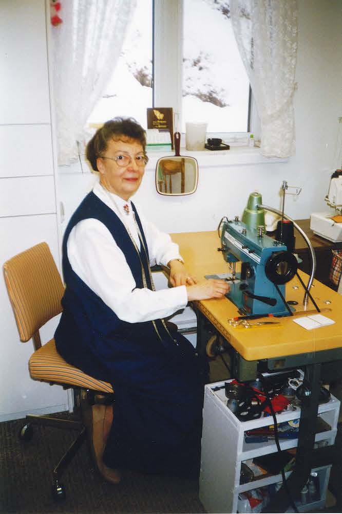 Photographie couleur de Marie-Rose Jalbert assise à sa machine à coudre, devant une fenêtre. Elle porte un ruban à mesurer autour du cou. On voit des accessoires de couture, un miroir et une lampe autour de son plan de travail.