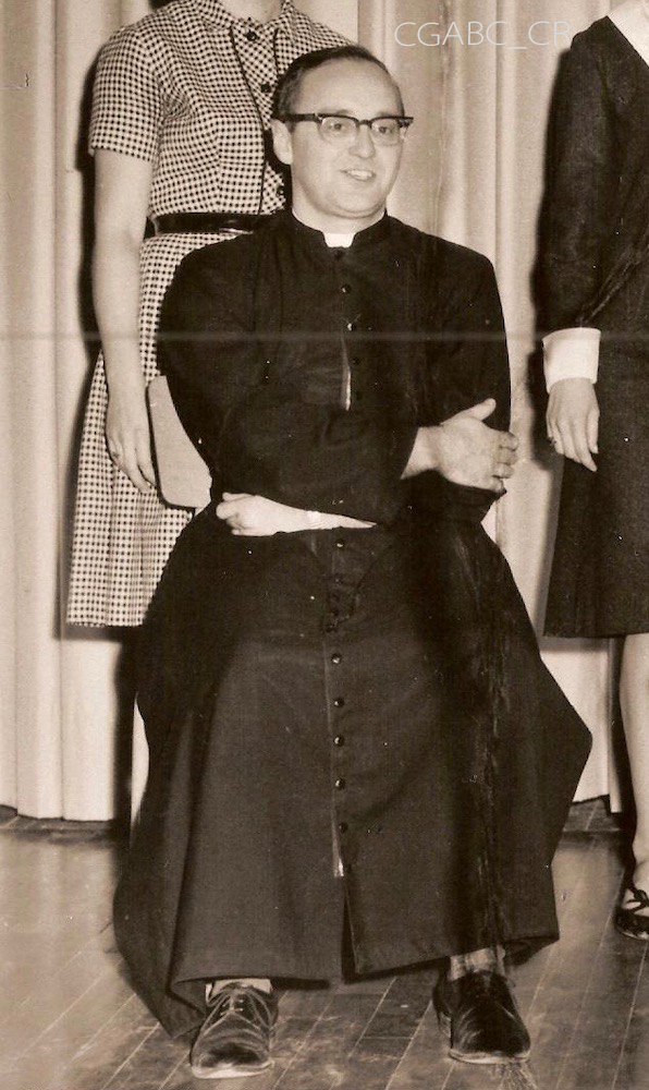 Photographie d’archives en noir et blanc montrant le vicaire Jean Moisan assis sur une chaise, les bras croisés. Il porte des lunettes à larges montures ainsi qu’une soutane et des souliers noirs. Deux femmes dont le visage n’est pas visible sont debout derrière lui.