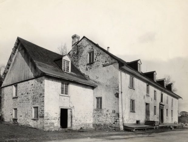 Photographie d’archives en noir et blanc montrant une vue éloignée de la façade et du profil gauche d’un édifice à pignons à trois étages. Le bâtiment est en pierre partiellement recouverte de chaux. Une annexe de plus petites dimensions se trouve à la gauche.