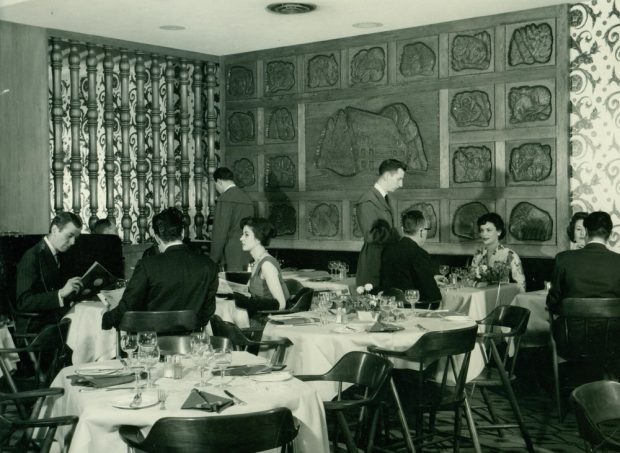 Photographie d’archives en noir et blanc montrant une salle à manger occupée par des serveurs et des clients attablés. Au fond de la salle, on aperçoit une murale en bois sculptée où un motif central est entouré de vingt-deux panneaux plus petits.
