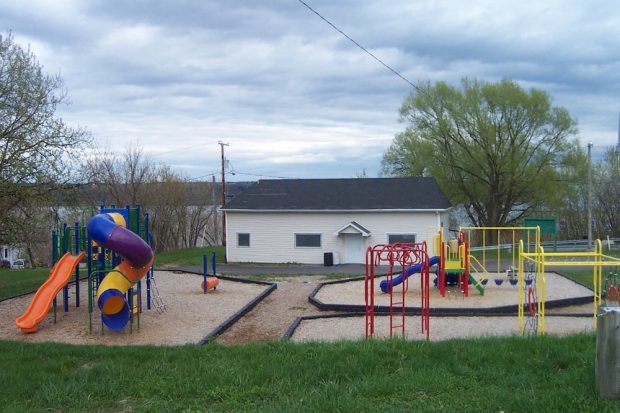 Photographie d’un parc où se trouvent un petit bâtiment blanc muni d’une porte et de trois fenêtres et, devant celui-ci, des modules de jeu pour les enfants. À gauche, on aperçoit un toboggan en colimaçon, une glissade et des jeux pour grimper et, à droite, des balançoires ainsi qu’une plus petite glissade. Le parc est entouré de gazon.