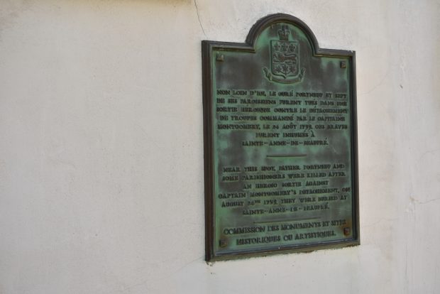 Photographie couleur d’une plaque en bronze où on peut lire : « Non loin d'ici, le curé Portneuf et sept de ses paroissiens furent tués dans une sortie héroïque contre le détachement de troupes commandé par le capitaine Montgomery, le 24 août 1759. Ces braves furent inhumés à Sainte-Anne-de-Beaupré. » La traduction anglaise suit. Au bas de la plaque, il est inscrit : Commission des monuments et sites historiques ou artistiques.