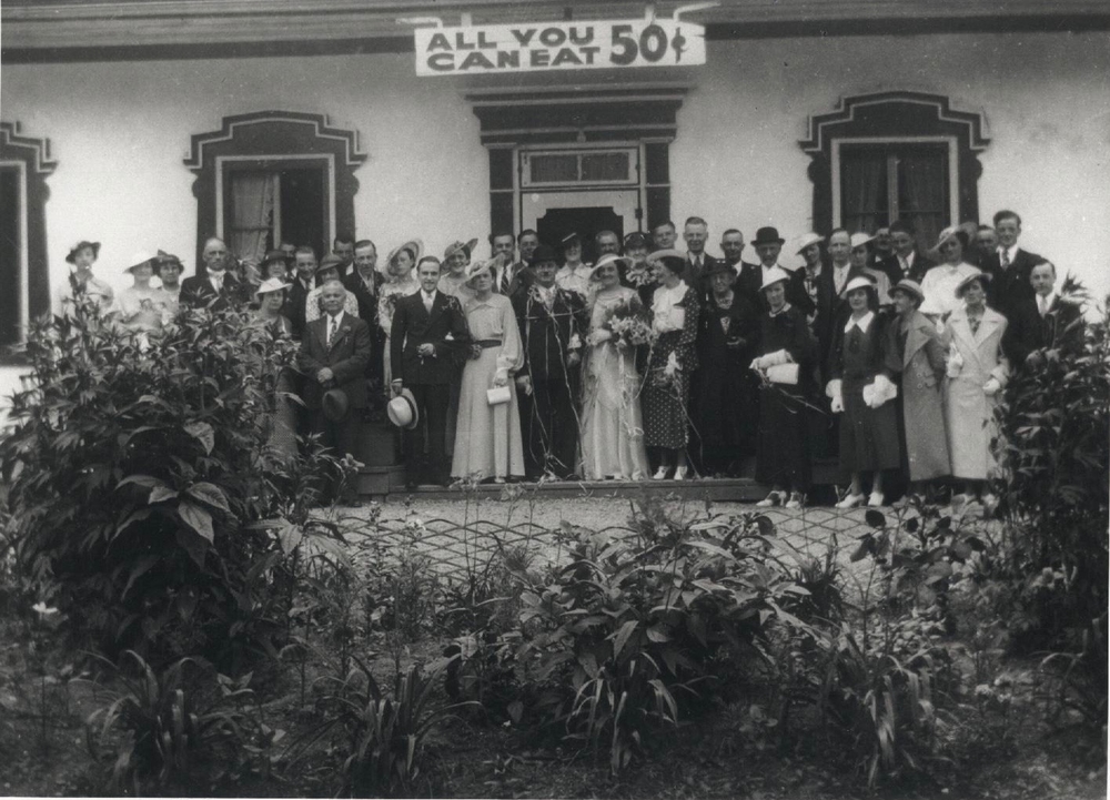 Photographie d’archives en noir et blanc montrant un couple de mariés entourés de leurs élégants invités devant l’auberge Baker. Tous regardent la caméra. Derrière le groupe, au-dessus de la porte, on voit une enseigne qui indique « ALL YOU CAN EAT 0,50$ ». L’avant-plan de la photographie est occupé par quelques plantes et arbustes.