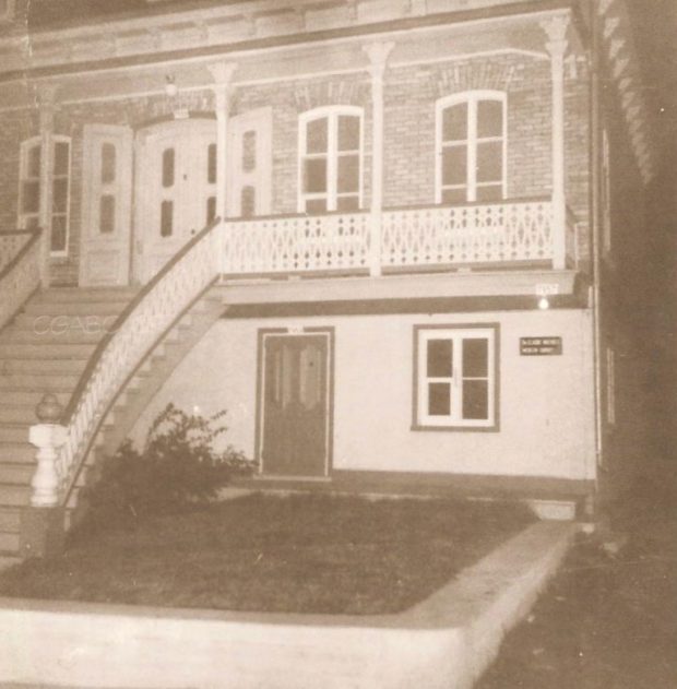 Photographie d’archives en noir et blanc d’une maison en briques divisée en deux étages. L’étage inférieur en crépi blanc est moins haut que l’étage supérieur. Un escalier menant à une galerie permet d’accéder à la porte principale du deuxième étage. De petites colonnes soutiennent un avant-toit qui couvre la galerie. Une plus petite porte adjacente à une fenêtre donne accès à l’étage inférieur.